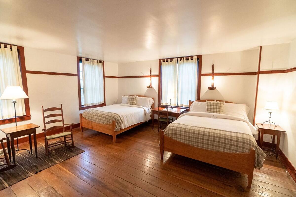 A hotel room with two single beds, a wooden floor, a desk with a chair, and a nightstand with a lamp.