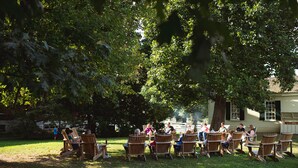 Un grupo de personas sentadas en sillas de madera al aire libre, bajo un árbol grande, con una casa al fondo.