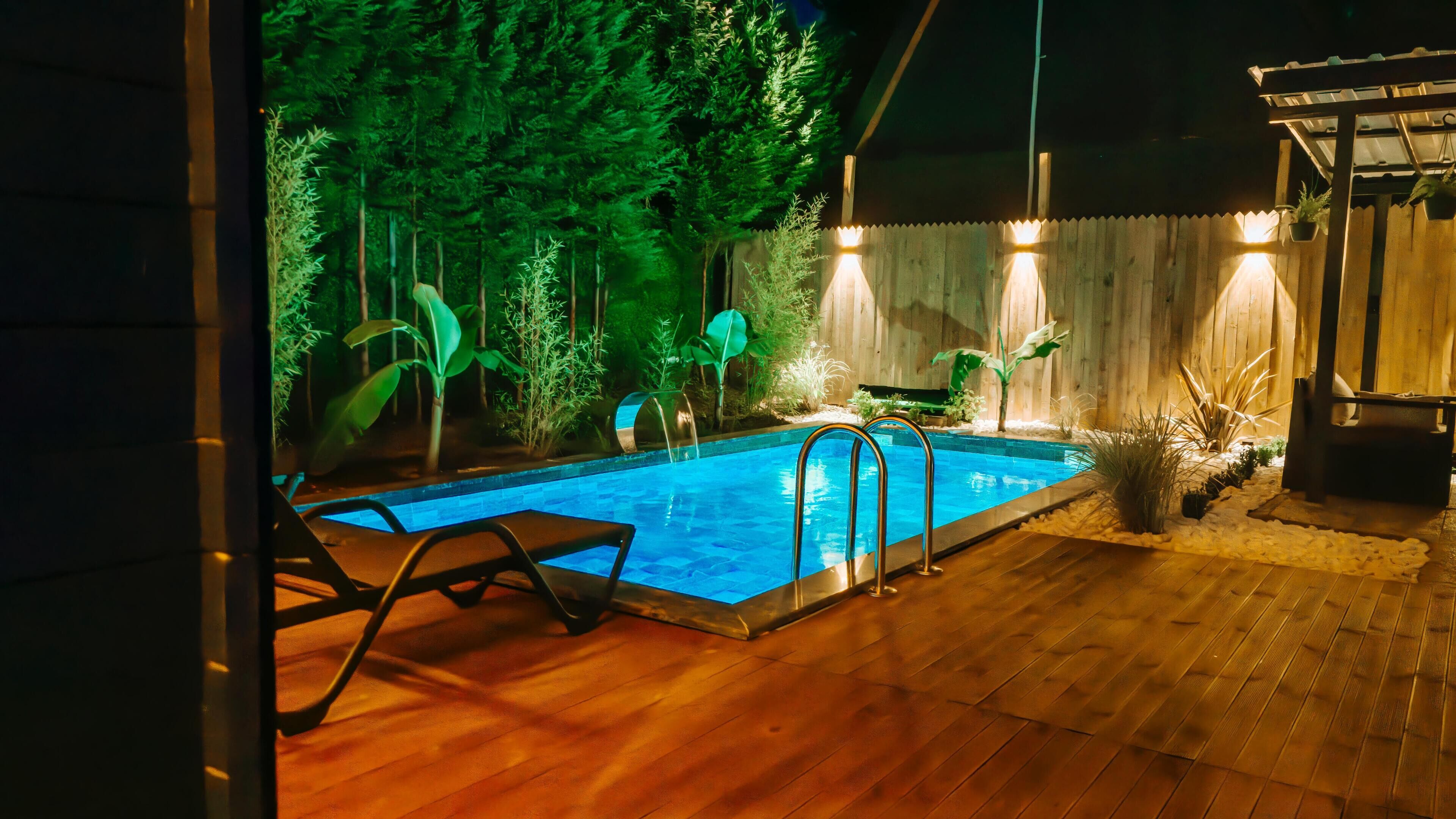 Piscina com deck de madeira e plantas ao redor, iluminada por iluminação ambiente.