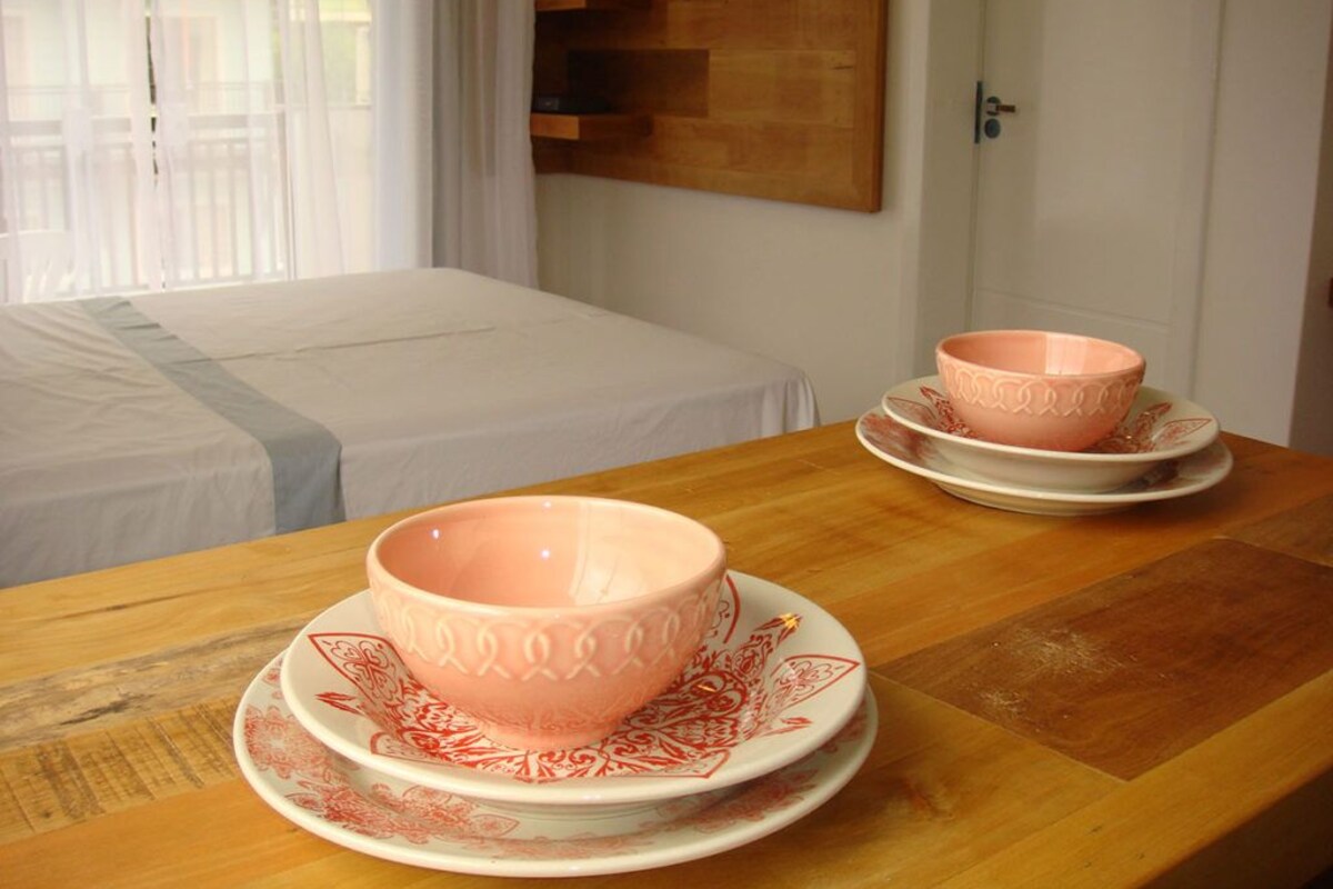 Une chambre d’hôtel avec une table en bois, deux bols roses assortis à leurs soucoupes, un lit recouvert d’une courtepointe blanche et une étagère en bois sur laquelle est posée une télévision.