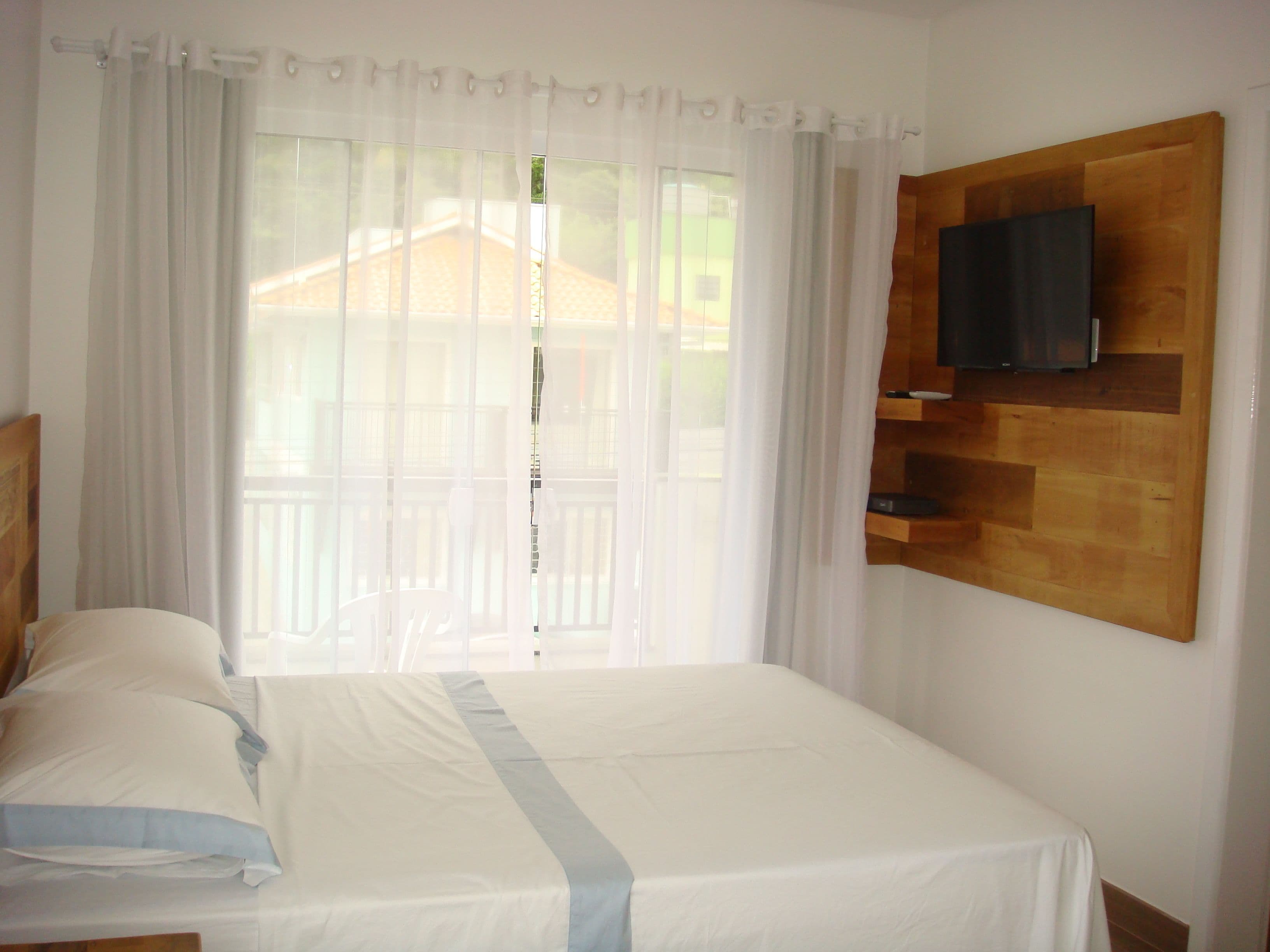 Habitación de hotel con cama, televisor montado en un panel de madera y balcón visible a través de cortinas translúcidas.