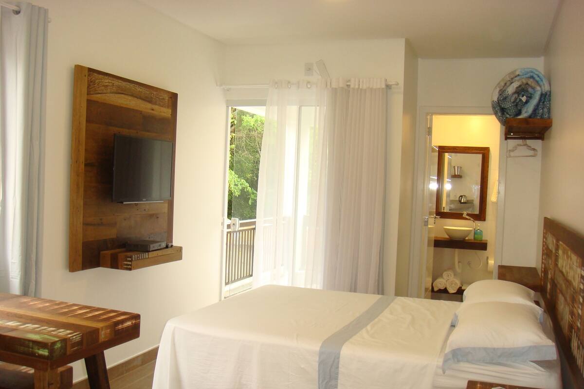 Habitación de hotel con cama, televisor montado en la pared, baño con lavabo y espejo, y balcón con cortinas.