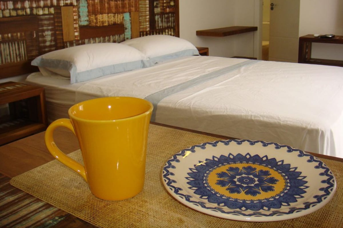 Un lit avec une tête de lit en bois, une tasse jaune et une assiette décorative posée sur un tapis tissé.