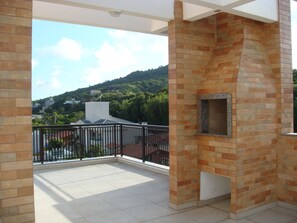 A balcony with a brick barbecue grill, a white ceiling, and a view of a green hillside with buildings. - Vento Solar Pousada (Bombinhas)