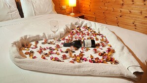 Una cama bien hecha con una botella de vino, copas y pétalos de flores esparcidos.