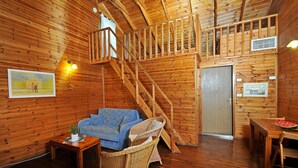 Interior de una cabaña de madera con una escalera, un sofá, sillas de mimbre, una mesa de comedor con sandía y cuadros en las paredes.