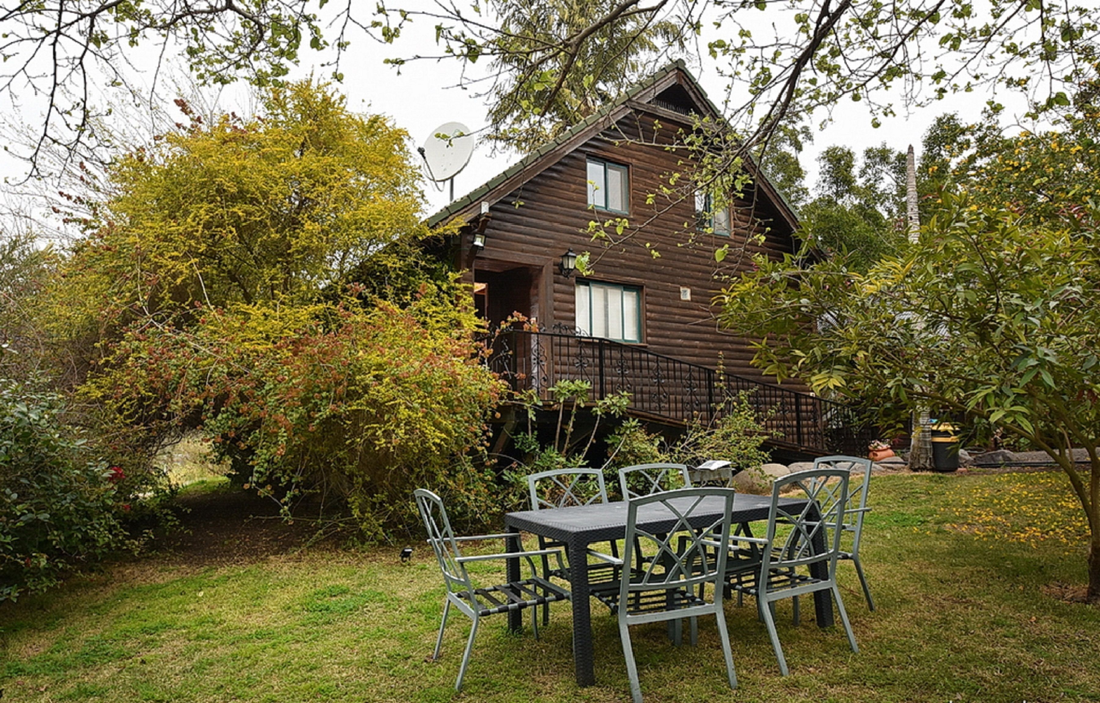 Uma cabana de madeira com varanda, rodeada por vegetação exuberante e uma área de refeições com cadeiras e mesa de metal.