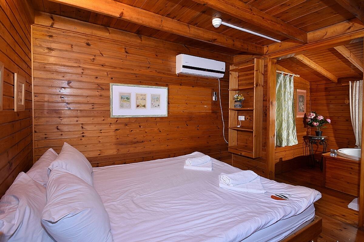 A wooden cabin room with a bed, a mounted air conditioner, framed pictures on the wall, and a bathroom with a sink and mirror.