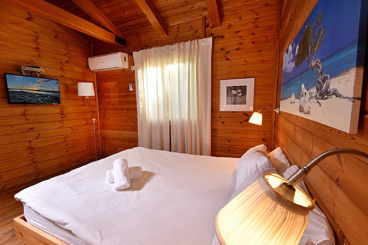 A wooden cabin room with a bed, a lamp, a painting, and a window with curtains.