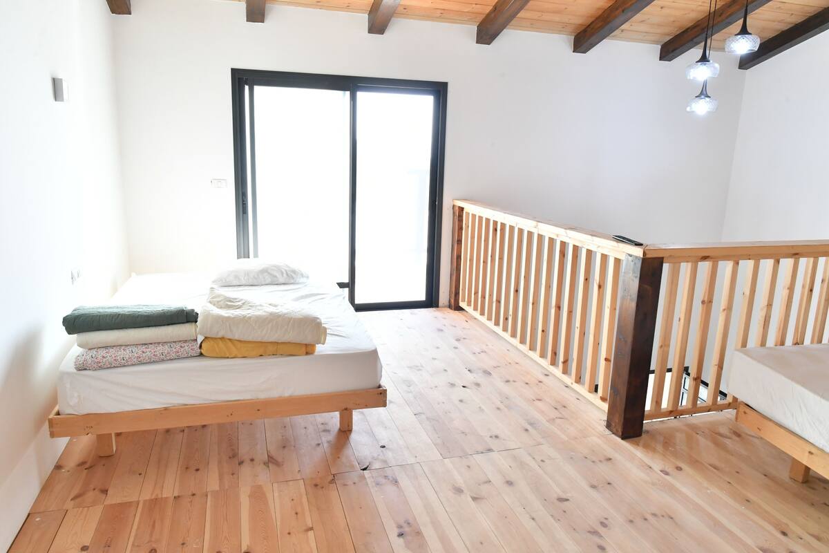 A room with a wooden floor, a bed with white bedding, a wooden railing, and a large window.