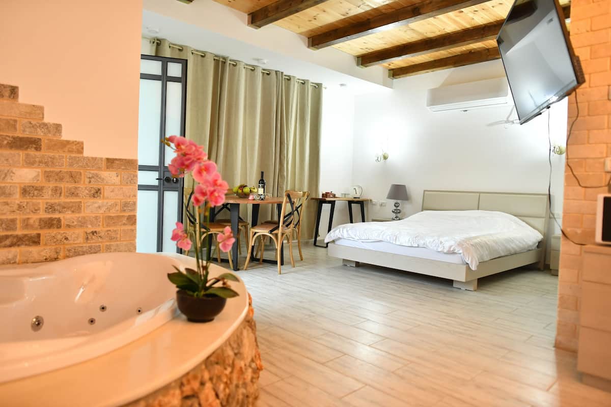 A bedroom with a bed, a desk, a chair, a TV, and a bathroom with a bathtub and a potted plant.