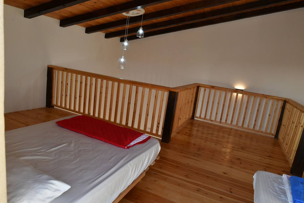 A bedroom with a wooden slatted headboard, a bed with white bedding and a red blanket, and a wooden floor.