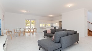 A spacious living area with a grey sofa, a dining table with chairs, a kitchen in the background, and a staircase leading to another floor.