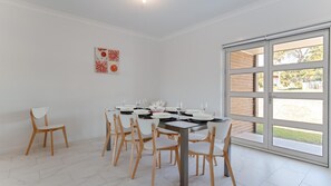Un coin repas avec une table dressée pour six personnes, entouré de chaises en bois, une grande fenêtre donnant sur un mur de briques et de la verdure, et une œuvre d’art accrochée au mur.