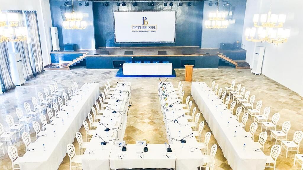 A conference room with rows of tables and chairs, a stage with a screen displaying 'PETIT BRUSSEL HOTEL - RESTAURANT - BEACH', and a chandelier.