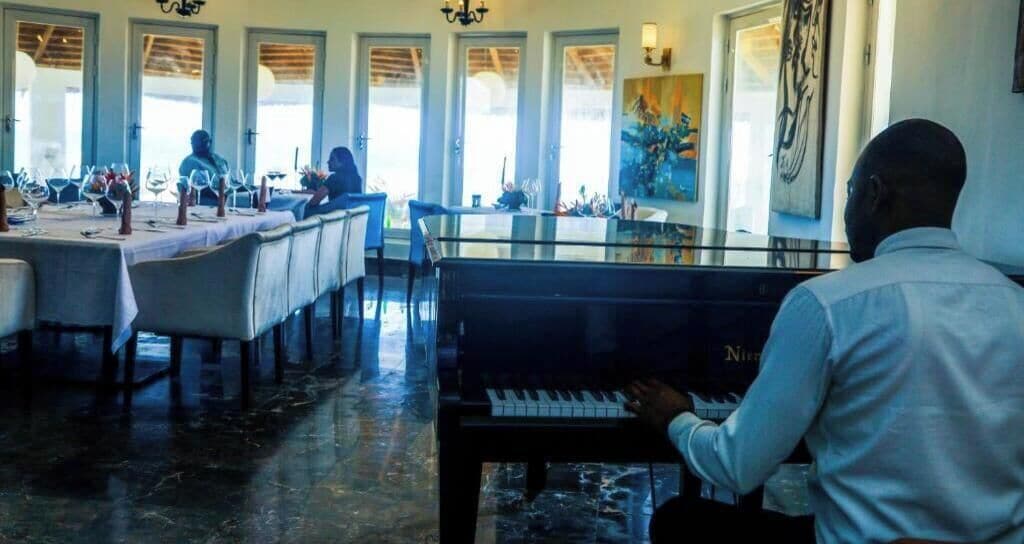 Un comedor con mesas dispuestas para una comida, un piano y ventanas amplias.