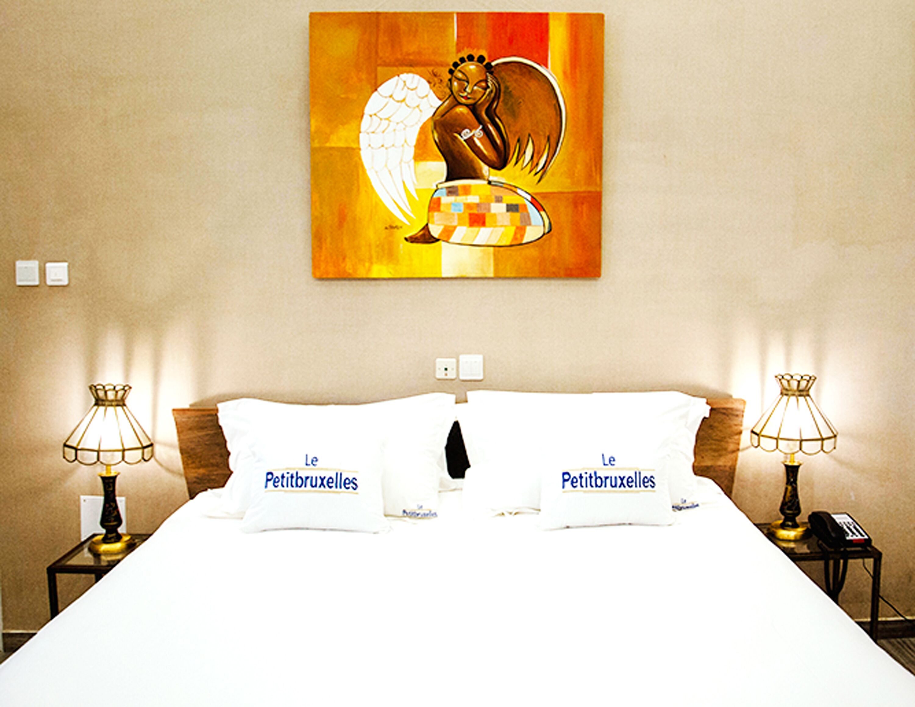Habitación de hotel con cama, dos lámparas de noche, un cuadro colgado en la pared y dos almohadas con el texto "Le Petitbruxelles".