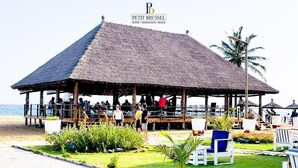 Un pabellón frente a la playa con techo de paja, rodeado de sillas de descanso y un pequeño jardín.