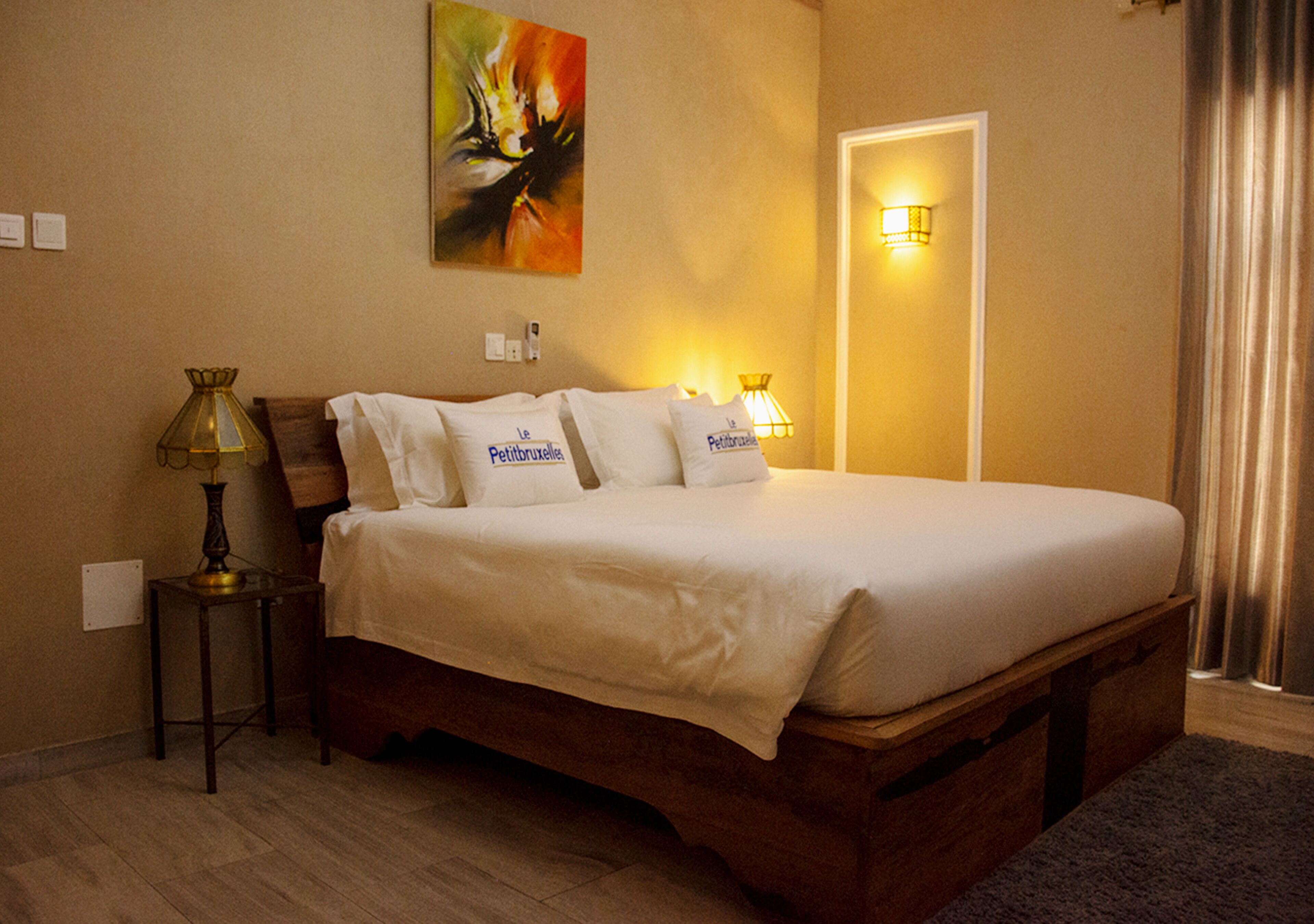 A neatly made bed with white linens and pillows, a bedside lamp, a painting on the wall, and a door with a light switch.