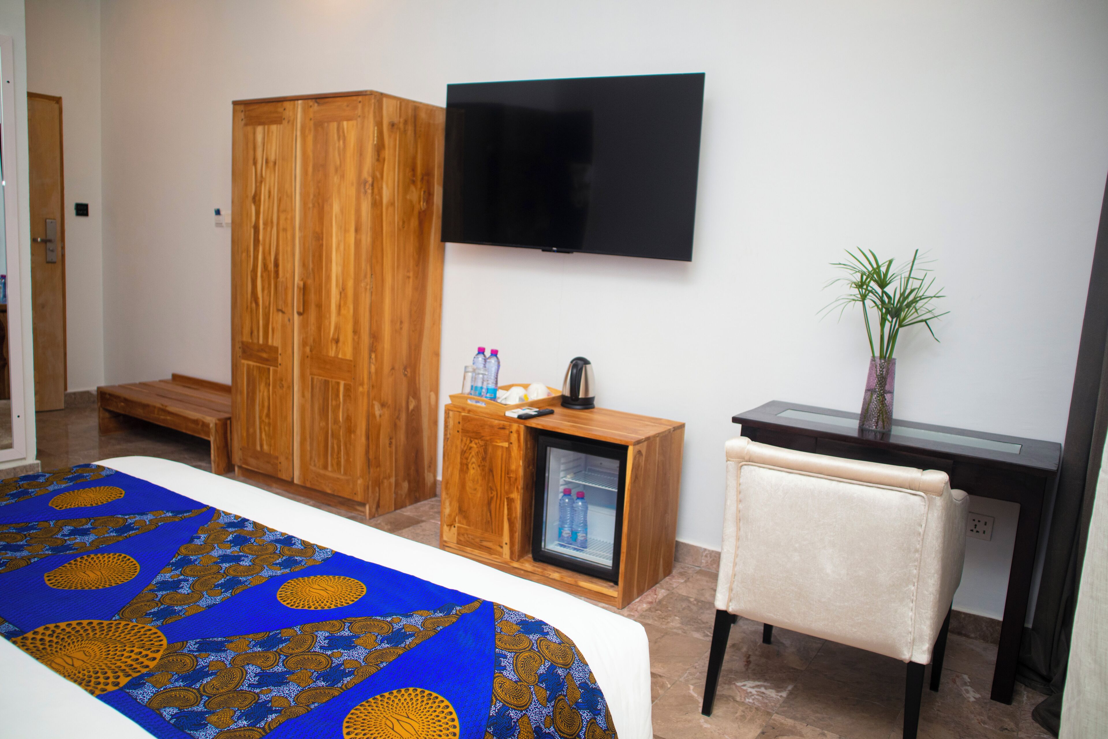 A hotel room with a bed, a television, a wooden wardrobe, a small refrigerator, a desk, and a chair.