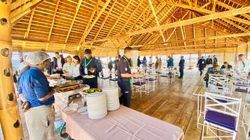 A covered dining area with tables and chairs, a high wooden ceiling, and a buffet setup with food trays.