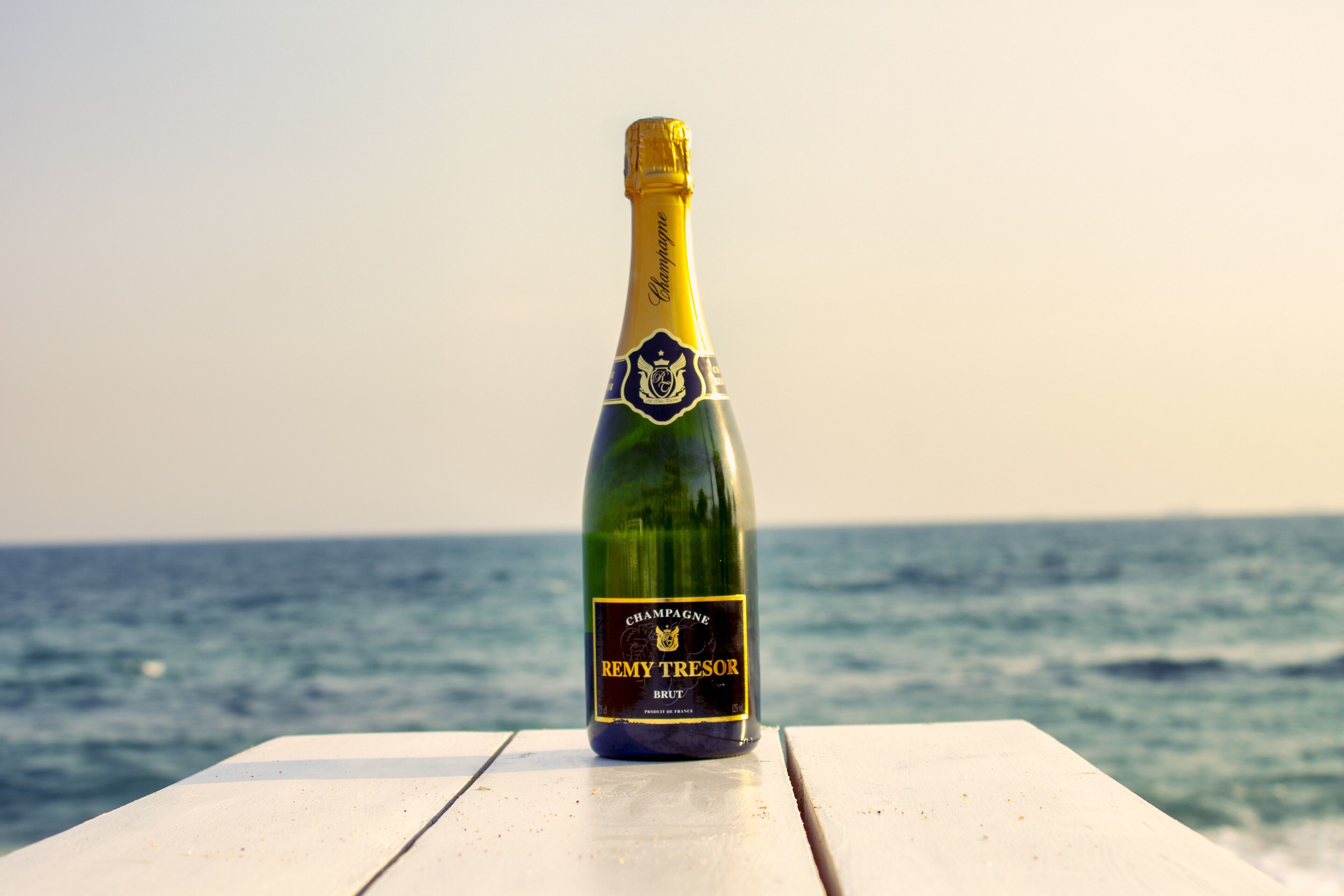 A bottle of Remy Tresor Brut champagne on a white wooden table by the sea.