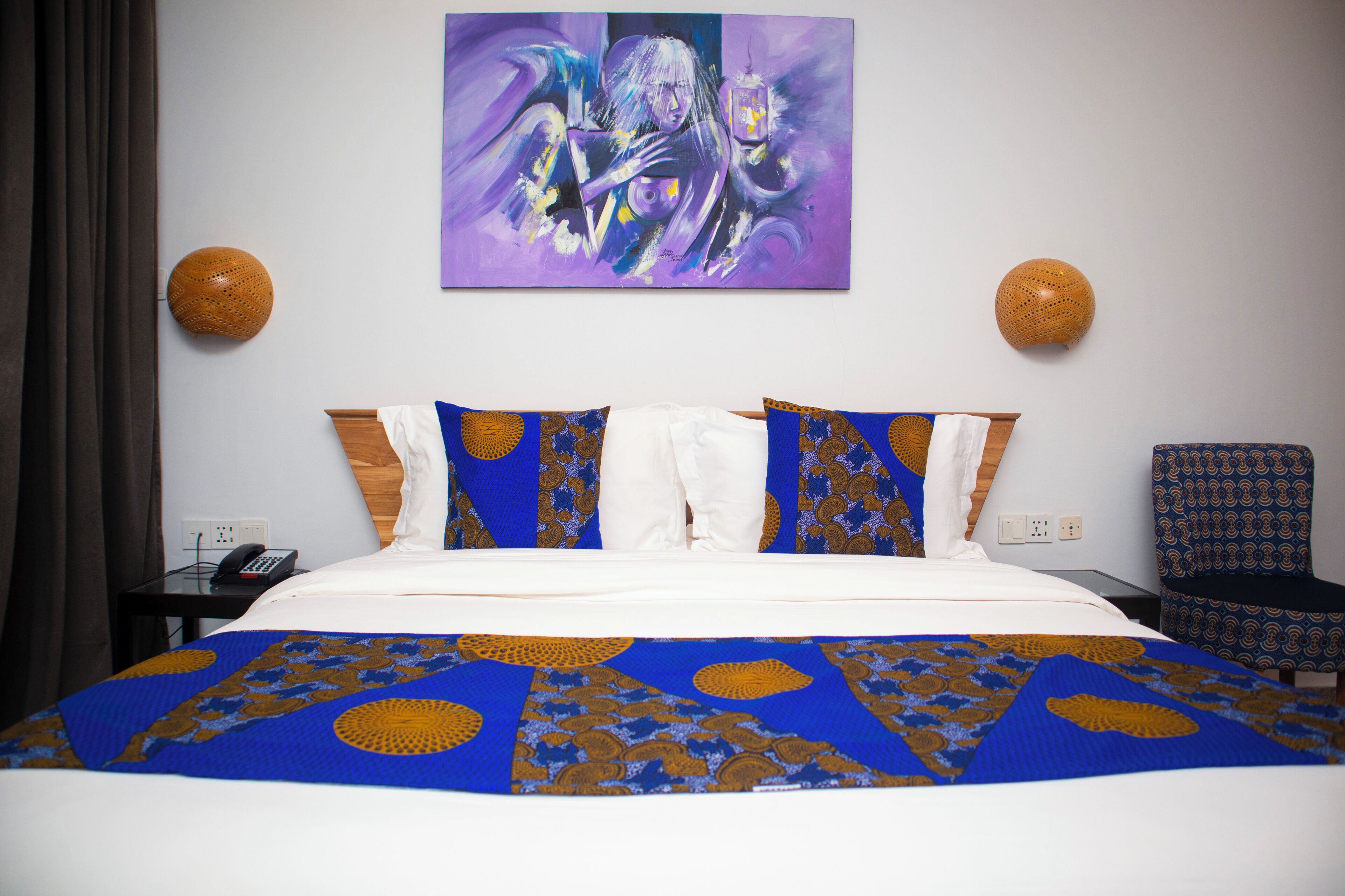 A hotel room with a bed, a painting on the wall, two wall sconces, a chair, and a telephone on a nightstand.