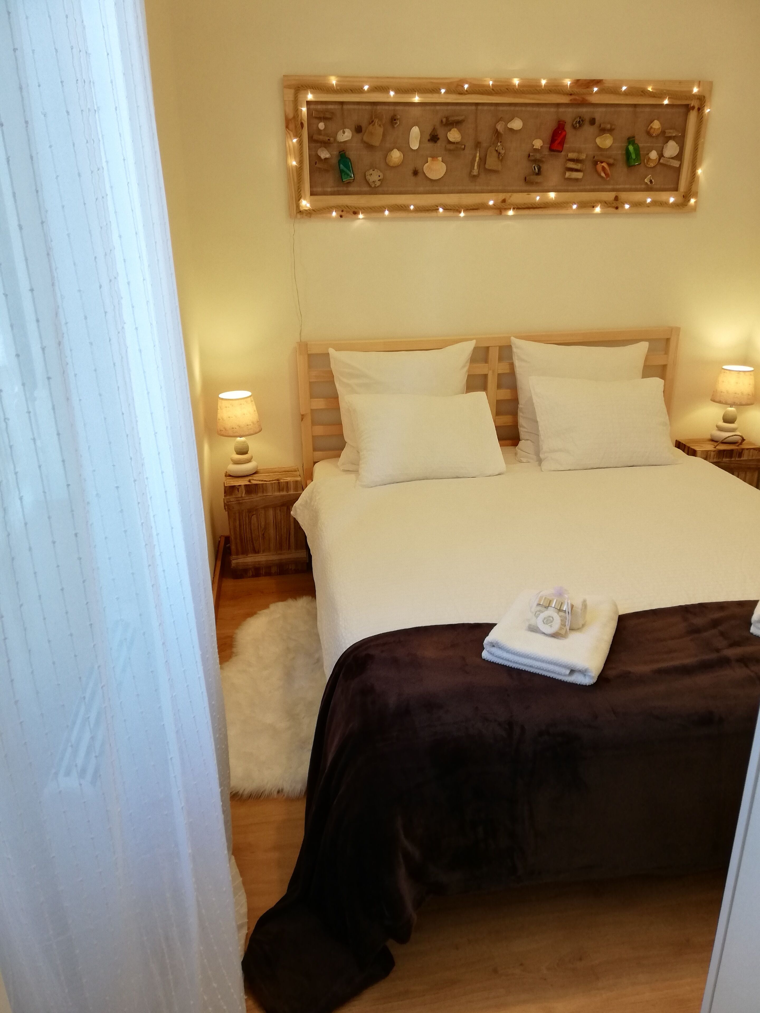 A neatly made bed with white linens and a dark brown bedspread, flanked by two bedside tables with lamps, a wooden headboard, and a wall-mounted lighted display of seashells and other items.