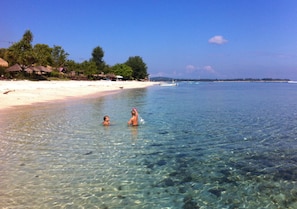 Ein Strand mit klarem, flachem Wasser, einem Sandufer und ein paar Booten in der Ferne.
