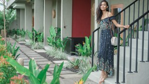 Una mujer con un vestido floral parada en una escalera con barandilla, rodeada de follaje y un edificio con puerta roja.
