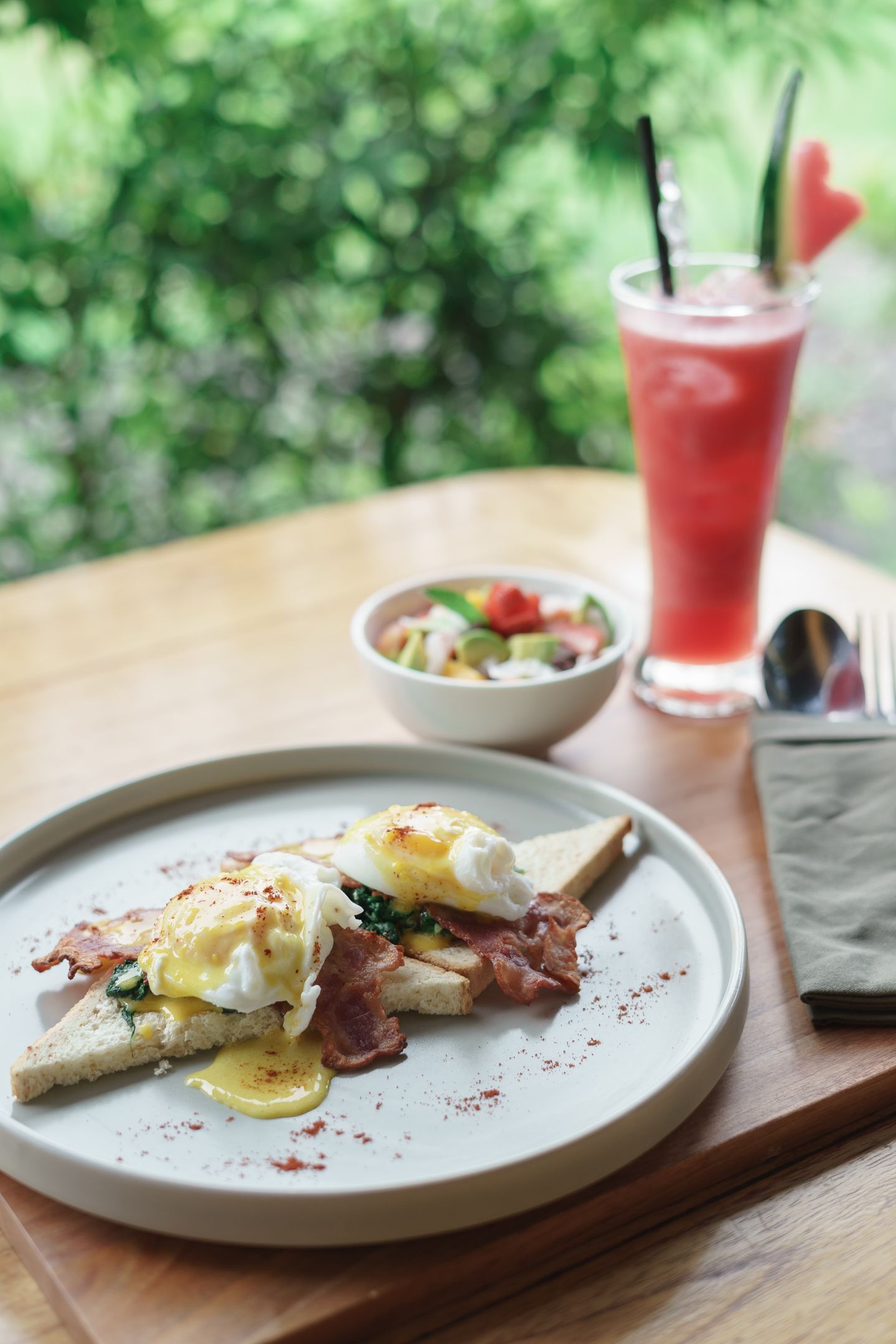 A breakfast plate with eggs, bacon, and toast, a bowl of salad, and a glass of red drink on a wooden table with a green background.