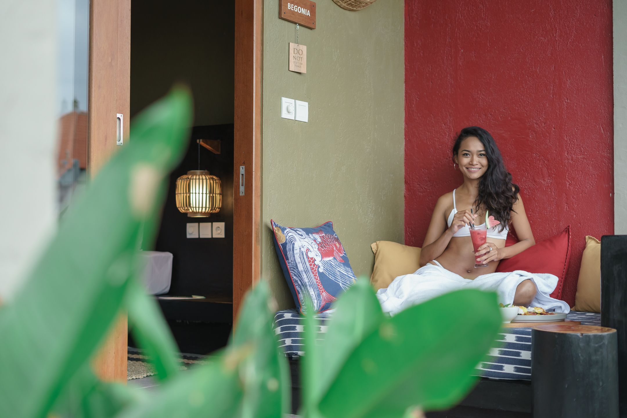 Una mujer sentada en un sofá con una bebida, una pared roja y un cartel que dice "BEGONIA" al fondo.