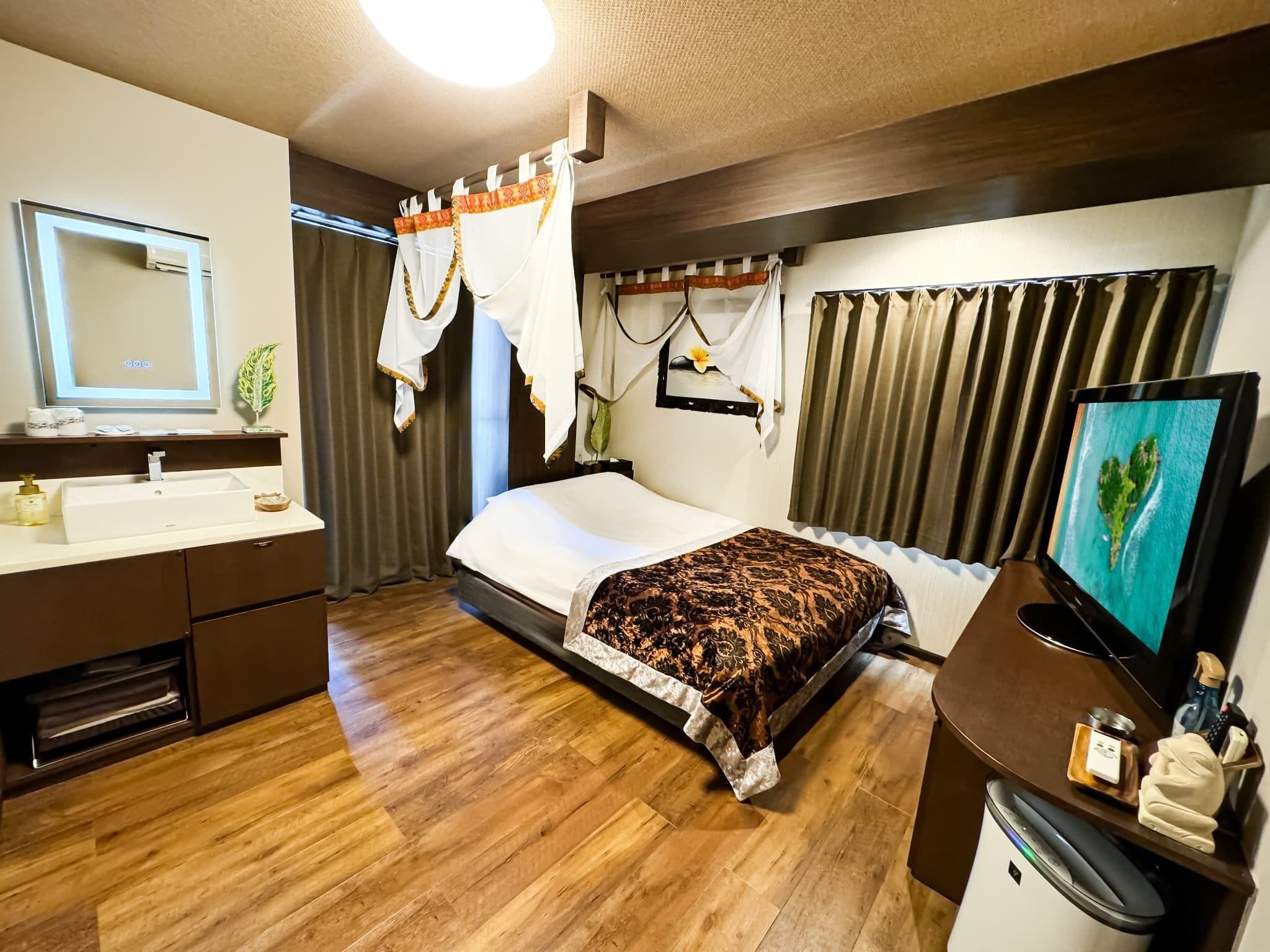 Una habitación de hotel con piso de madera, una cama con cubrecama estampado, una mesita de noche con televisor, un baño con lavamanos y espejo, y cortinas.
