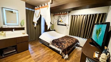 Una habitación de hotel con piso de madera, una cama con cubrecama estampado, una mesita de noche con televisor, un baño con lavamanos y espejo, y cortinas.