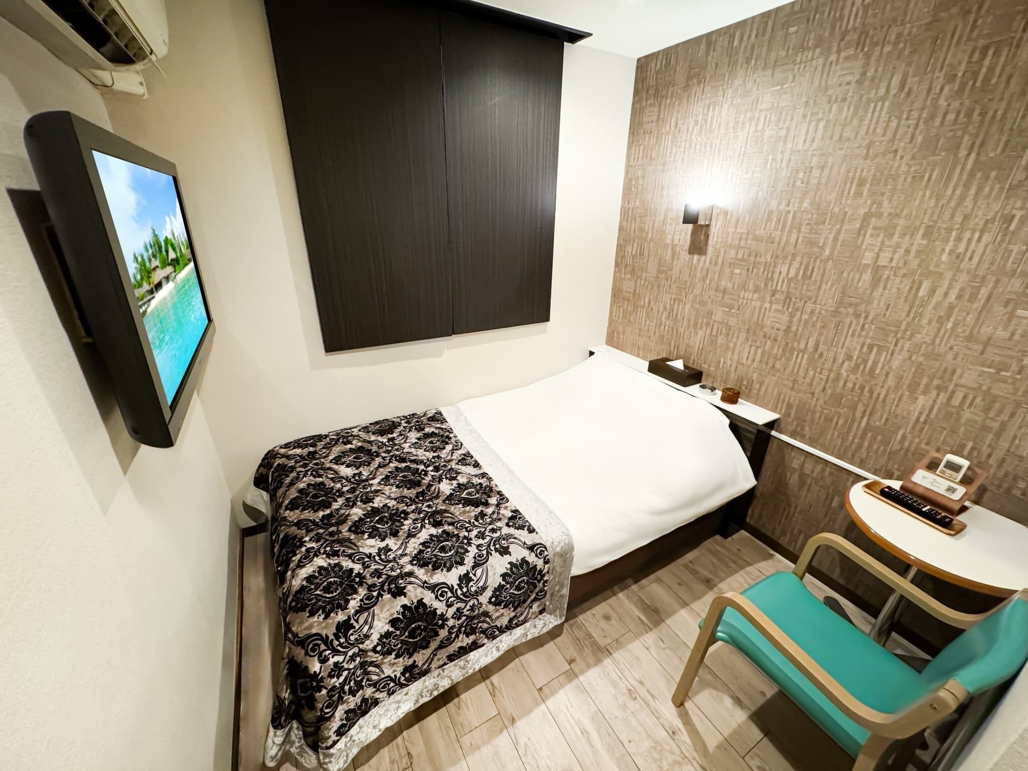 A hotel room with a bed, a TV mounted on the wall, a small table with a chair, and a wooden floor.