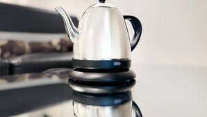 Een roestvrijstalen theepot met zwart handvat en deksel op een reflecterend oppervlak.
