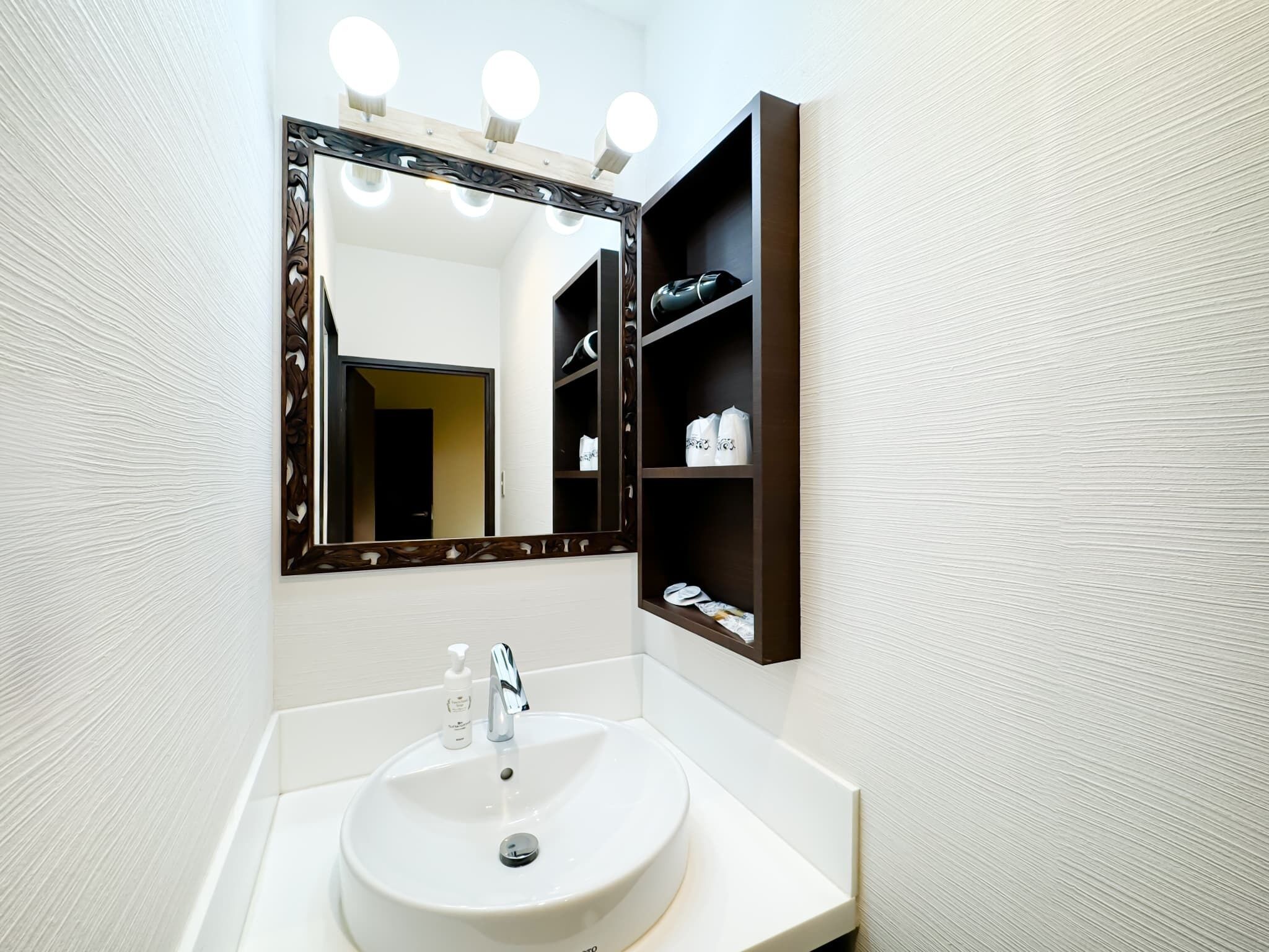 A bathroom with a white sink, a mirror with lights, and a shelf with toiletries.