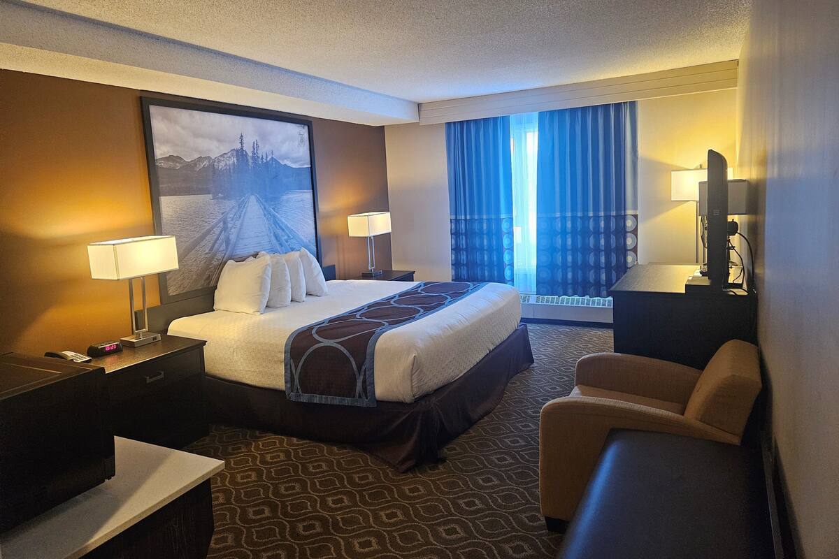 A hotel room with a bed, two bedside tables, a desk, a chair, a large window with curtains, and a wall-mounted TV.