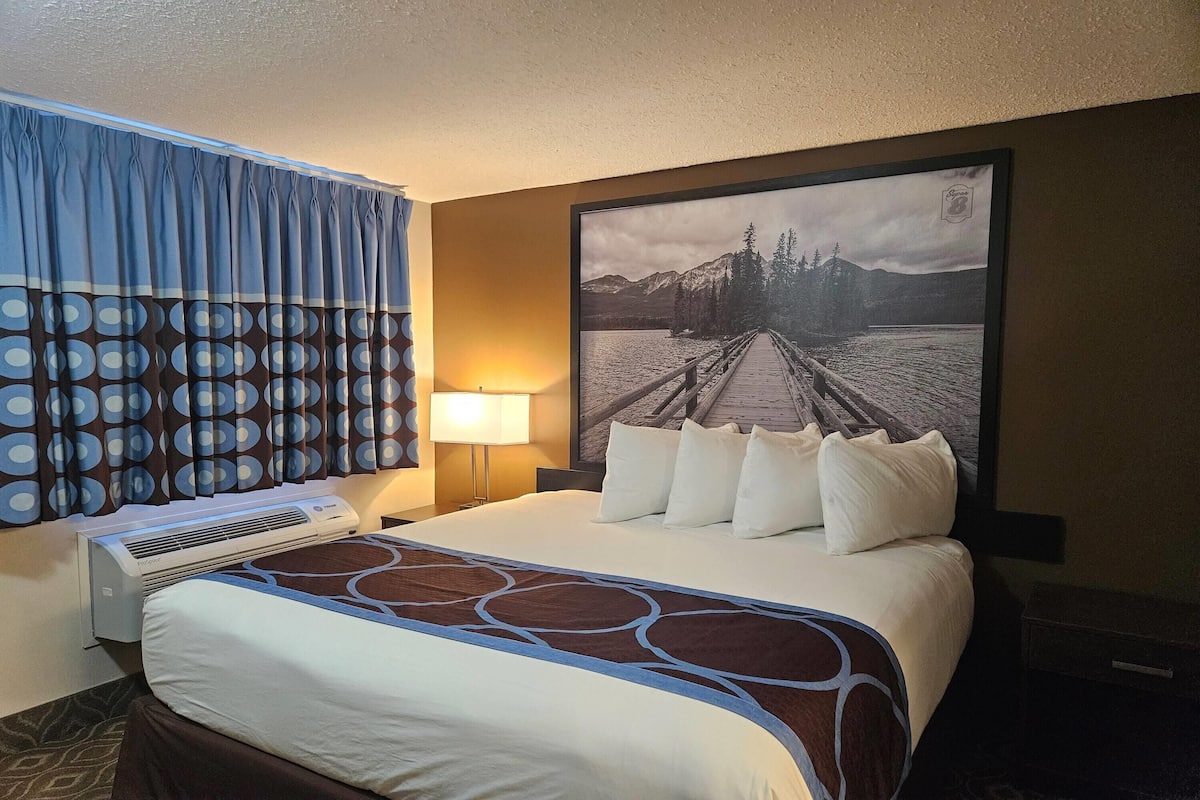 A hotel room with a bed, a large window with curtains, a wall-mounted air conditioner, and a framed picture of a bridge over water.
