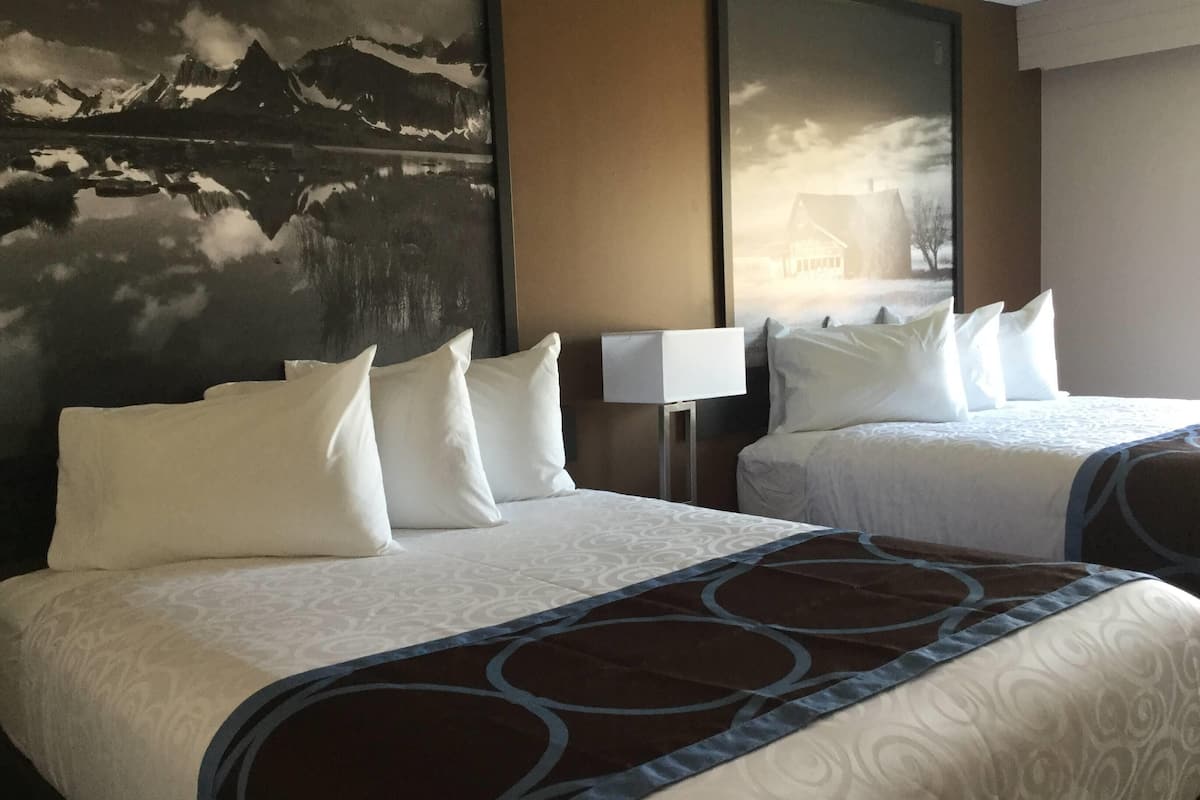 A hotel room with two beds, each with white linens and pillows, a brown patterned throw blanket, and a large wall painting of a mountain landscape.