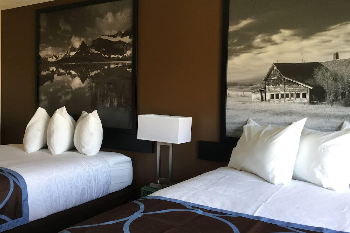 A hotel room with two beds, a nightstand, a lamp, and two large landscape paintings on the walls.