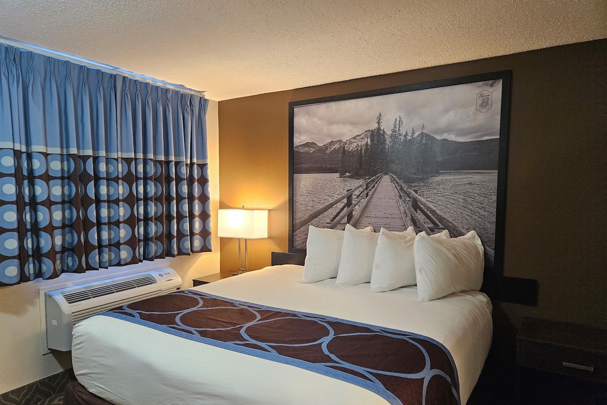 A hotel room with a bed, a desk, a lamp, and a large framed picture of a lake and mountains.