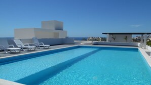 Une piscine rectangulaire à l’eau d’un bleu cristallin, entourée de chaises longues blanches et bordée d’un bâtiment blanc.