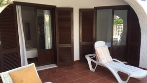Un espacio de estar al aire libre cubierto con dos sillones blancos, uno con cojín color beige, y una silla de mimbre con cojín blanco.