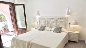 Una cama doble con colcha blanca y dos almohadones a juego, flanqueada por mesitas de noche con lámparas, en una habitación con suelo de madera y un cabecero decorativo.
