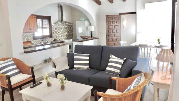 Una sala de estar acogedora con un sofá oscuro, dos sillones de mimbre, una mesa de centro blanca y una cocina al fondo.