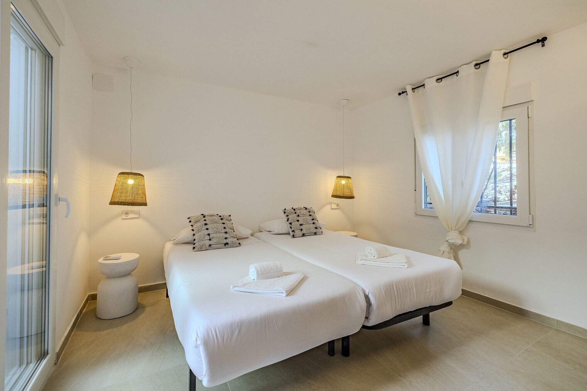 A clean, minimalist bedroom with two beds, bedside lamps, a window with curtains, and a toilet.
