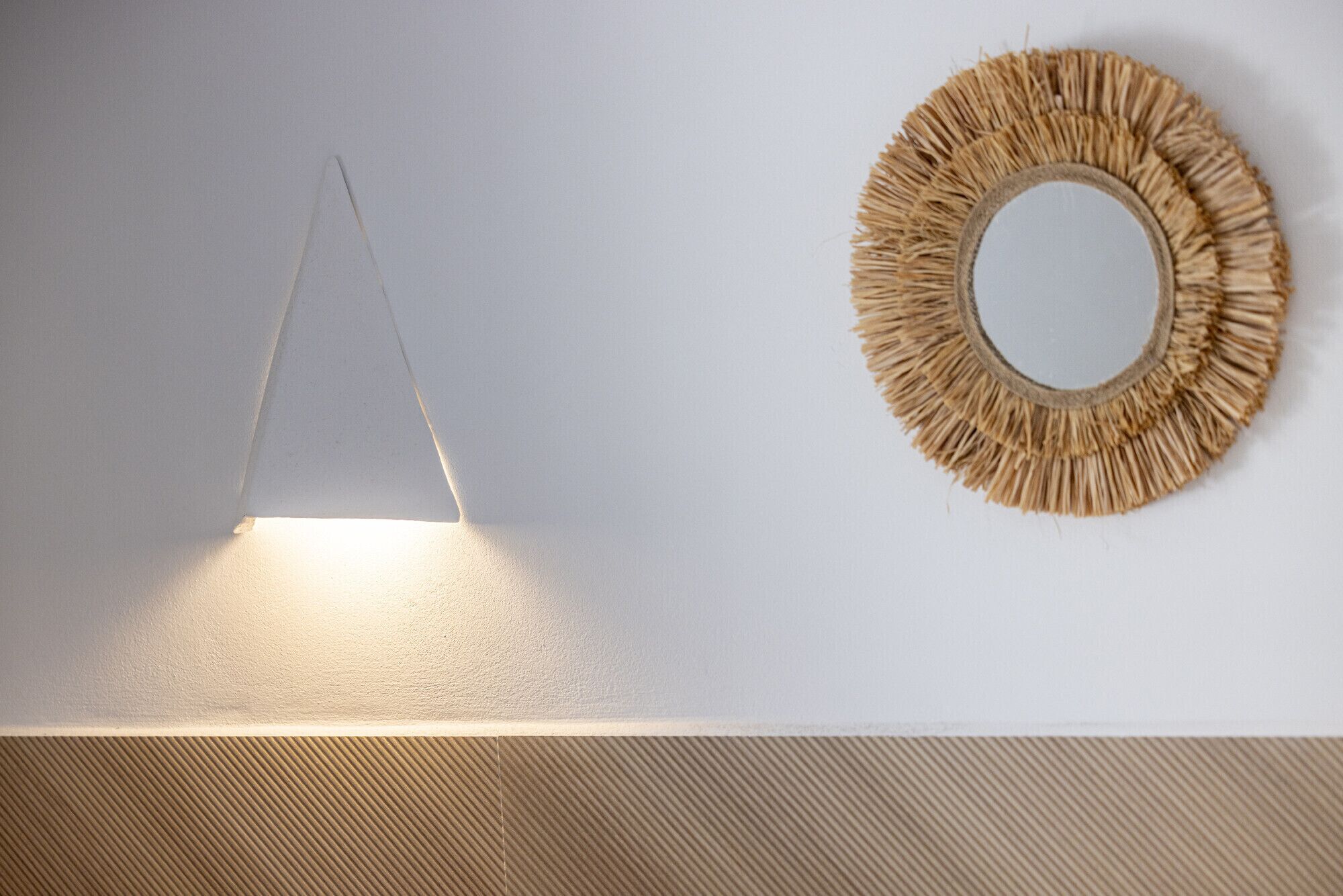 A wall with a hanging triangular light fixture and a circular woven mirror.