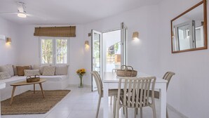Un salon moderne avec un canapé, une table basse et une table à manger avec des chaises. Il y a une fenêtre avec des stores, un ventilateur de plafond et un miroir au mur.