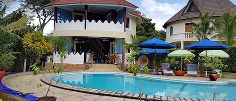 Zona de piscina con piscina, sombrillas y un edificio con tejado rojo.